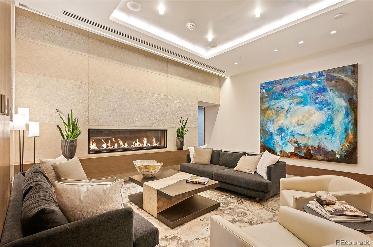 This is a modern living room featuring a linear fireplace set within a textured wall, complemented by contemporary furniture including a gray sofa and cream-colored armchairs. An abstract painting adds a splash of color, and the room is illuminated by recessed lighting and floor lamps, creating a warm and inviting atmosphere. The perspective is from a medium distance, showcasing the entire space.