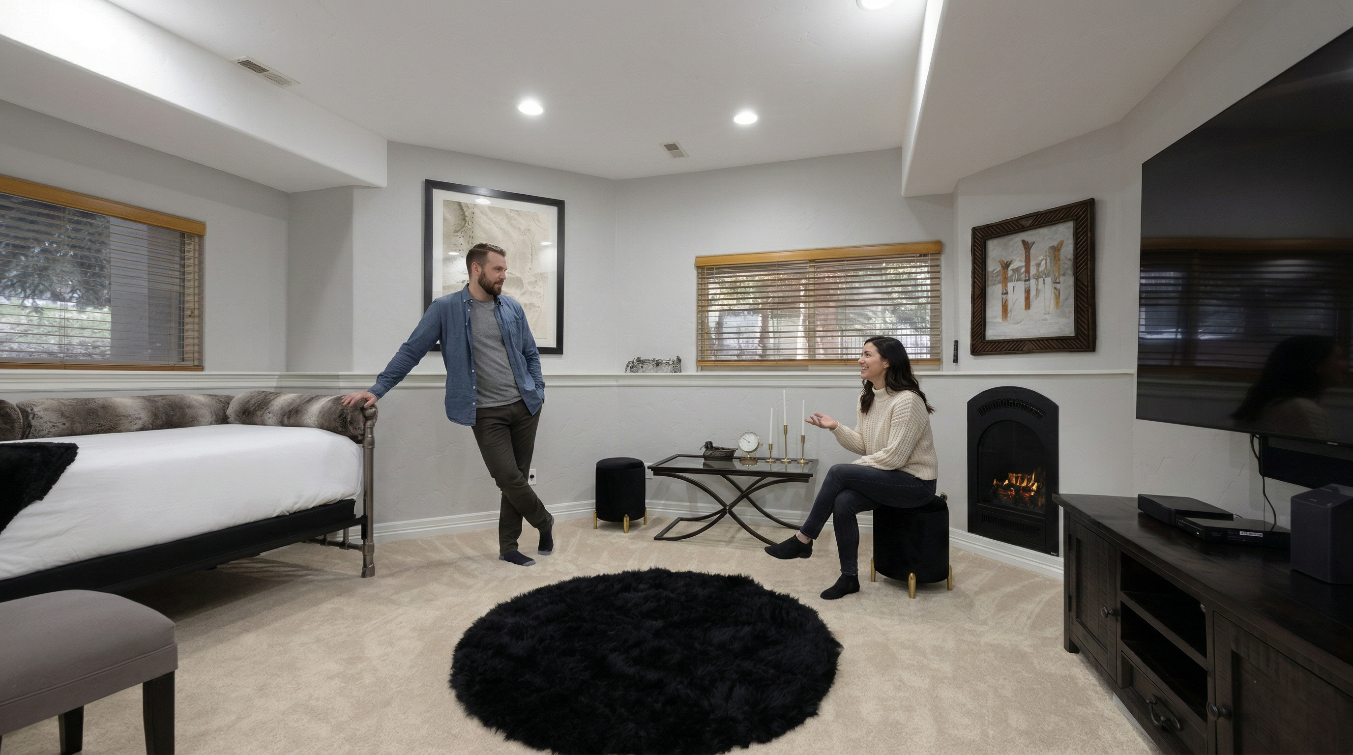 This cozy, finished basement features a neutral color palette with plush carpeting and a dedicated sleeping area with a daybed. The space is anchored by a corner fireplace, a sleek glass-top coffee table with black ottomans, and a large television, creating a comfortable lounge environment. The room is well-lit with recessed lighting and accented by tasteful wall art, offering a versatile space for relaxation or hosting guests.