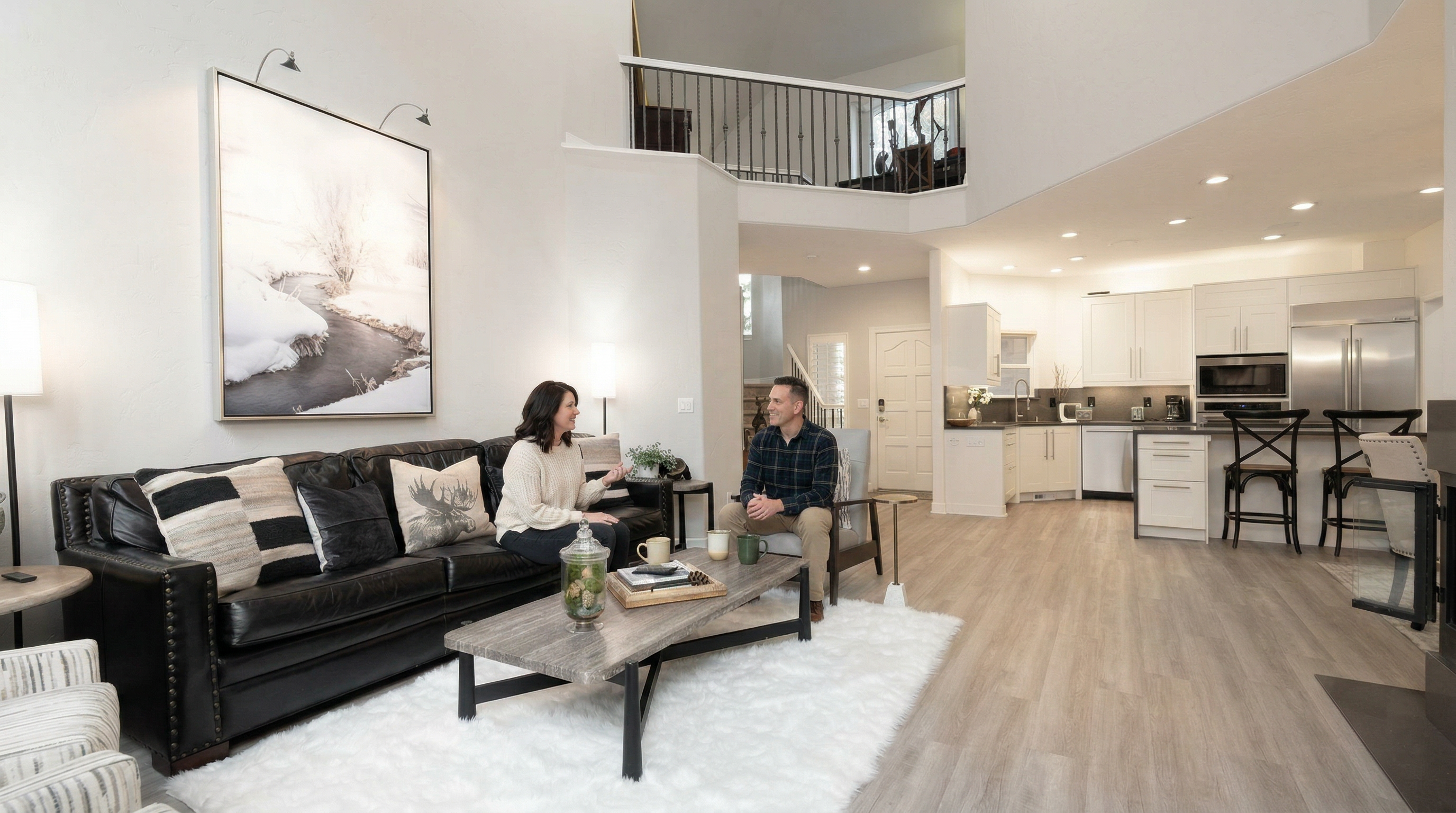 This spacious, open-concept living room features a high-contrast design with a large black leather sofa, a rustic wood coffee table, and a plush white area rug. The room flows seamlessly into a modern kitchen with white cabinetry and bar seating, all under a soaring vaulted ceiling that overlooks a lofted balcony. The bright, airy atmosphere is enhanced by neutral tones and contemporary decor, creating an inviting and sophisticated living space.