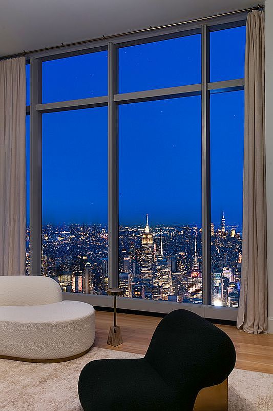 This sophisticated living space features floor-to-ceiling windows that frame a breathtaking, panoramic night view of the New York City skyline, including the iconic Empire State Building. The room is furnished with a minimalist, cream-colored curved sofa and a sleek black accent chair, complemented by a small, elegant side table. The combination of the plush, neutral-toned rug and the dramatic urban backdrop creates an atmosphere of modern luxury and high-rise exclusivity.
