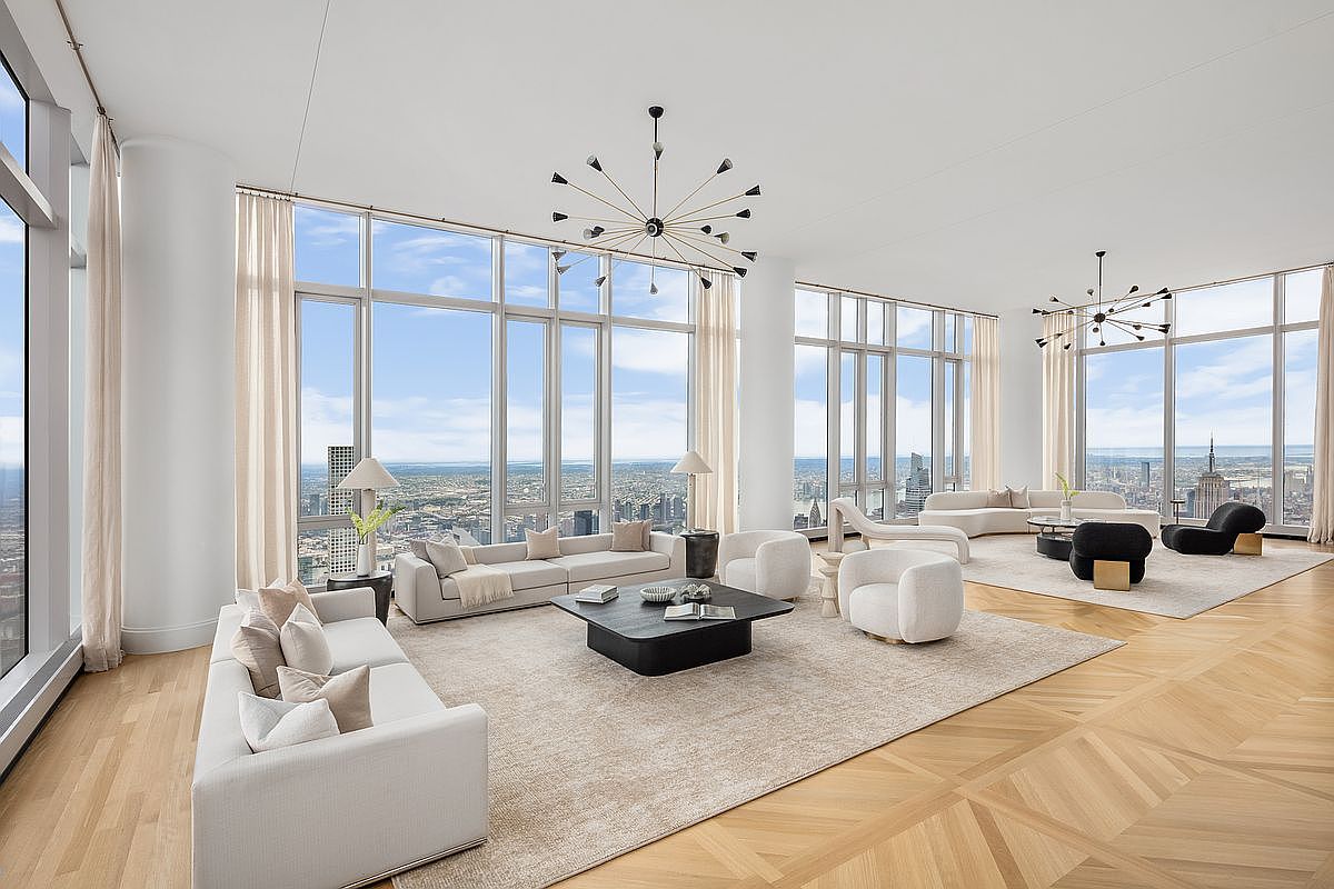 This expansive, high-floor living room features floor-to-ceiling windows that offer panoramic city views, including the Empire State Building. The space is elegantly furnished with two distinct seating areas, featuring plush white sofas, modern armchairs, and minimalist coffee tables set atop neutral area rugs. The room is anchored by light-toned herringbone wood flooring and striking sputnik-style chandeliers, creating an atmosphere of sophisticated, airy luxury.
