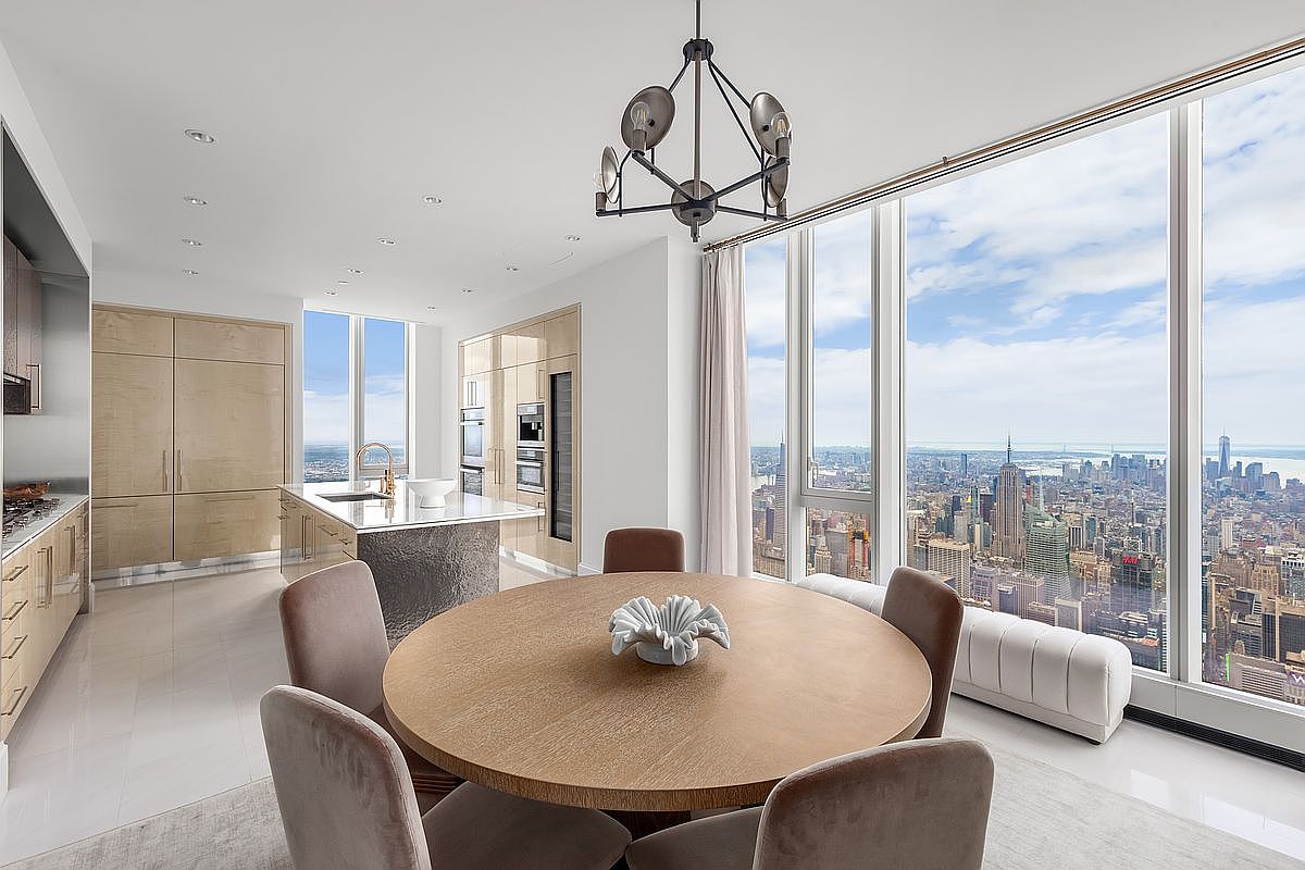 This sophisticated dining area features a round wooden table surrounded by plush, neutral-toned chairs, positioned beneath a modern geometric chandelier. The space seamlessly integrates with a sleek, high-end kitchen, while floor-to-ceiling windows offer a breathtaking, panoramic view of the New York City skyline. The overall aesthetic is one of refined luxury, characterized by clean lines, light wood cabinetry, and an airy, open-concept layout.
