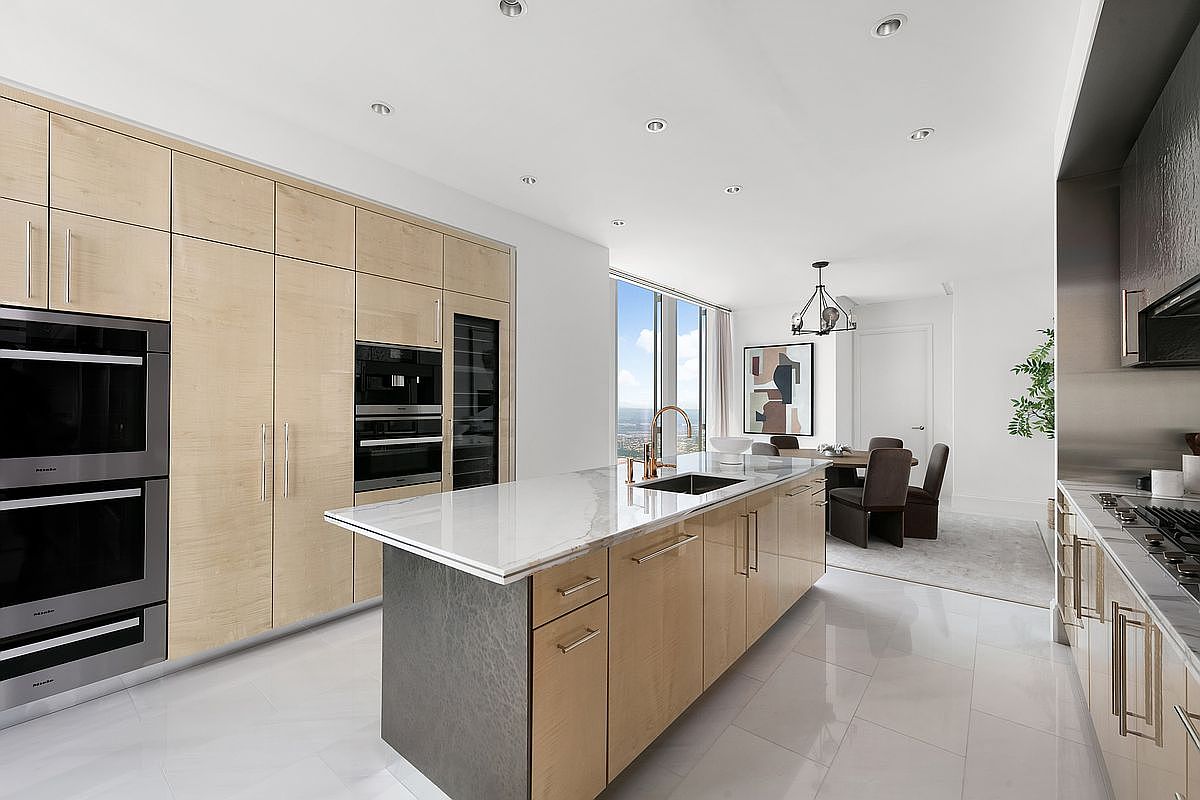This sleek, modern kitchen features light wood cabinetry, a large marble-topped island with a built-in sink, and high-end stainless steel appliances. The open-concept design flows seamlessly into a dining area, with floor-to-ceiling windows providing a bright, airy atmosphere and expansive city views. The polished tile flooring and minimalist aesthetic create a sophisticated and luxurious living space.