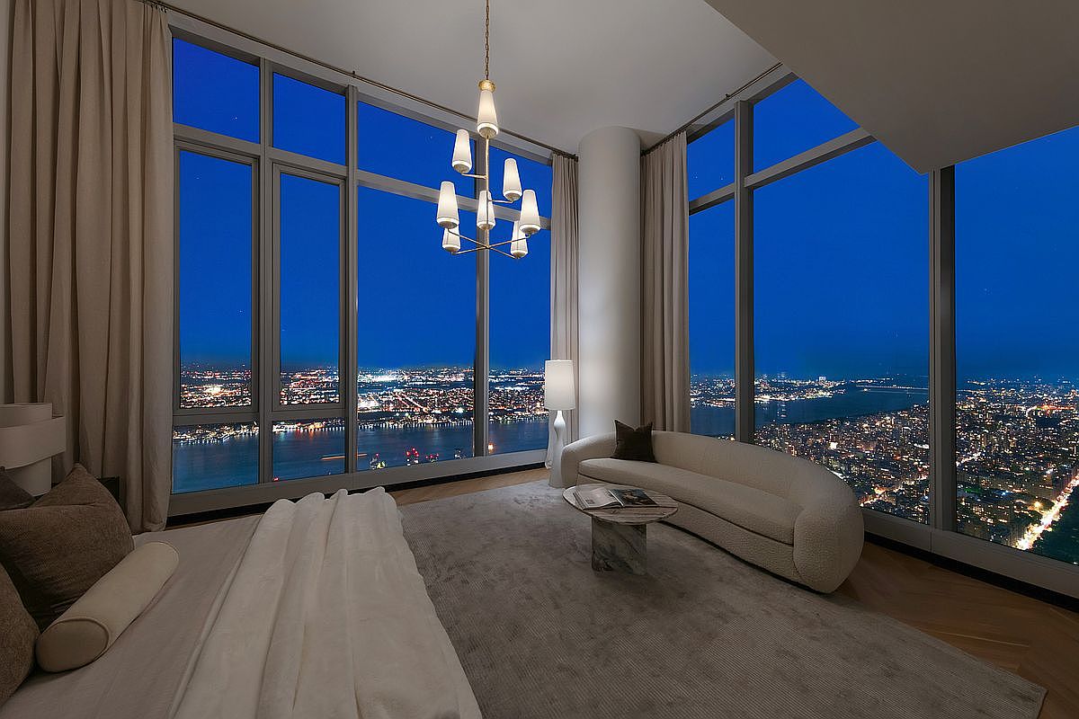 This luxurious primary bedroom features floor-to-ceiling windows that offer a breathtaking, panoramic night view of the city skyline and river. The room is elegantly furnished with a plush, curved sofa, a marble-topped coffee table, and a sophisticated chandelier, all set against a neutral, minimalist color palette. The perspective captures the expansive scale of the space, emphasizing the seamless blend of high-end interior design and dramatic urban scenery.