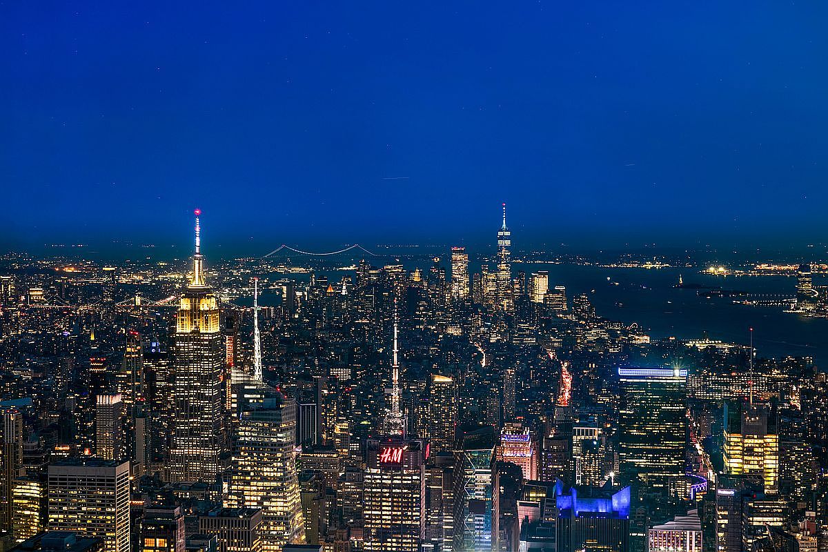 This stunning aerial night shot captures the iconic New York City skyline, dominated by the illuminated Empire State Building and the distant One World Trade Center. The dense urban landscape is a tapestry of glowing windows and street lights, creating a vibrant and sophisticated metropolitan atmosphere. The perspective is high-altitude, offering a cinematic, sweeping view of the city's architectural grandeur and bustling energy.