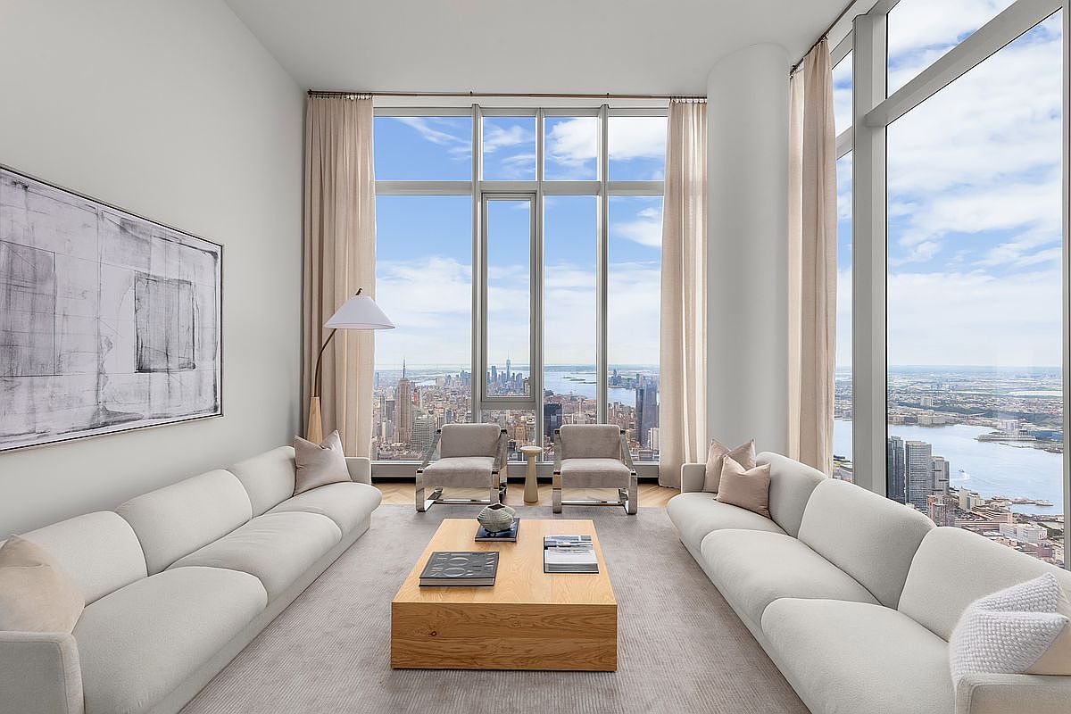 This sophisticated, high-rise living room features a symmetrical layout with two plush, light-colored sofas facing each other across a minimalist wooden coffee table. Floor-to-ceiling windows provide a breathtaking, panoramic view of the New York City skyline, bathing the room in natural light. The space is elegantly appointed with neutral tones, contemporary art, and sleek accent chairs, creating an atmosphere of refined luxury and urban serenity.
