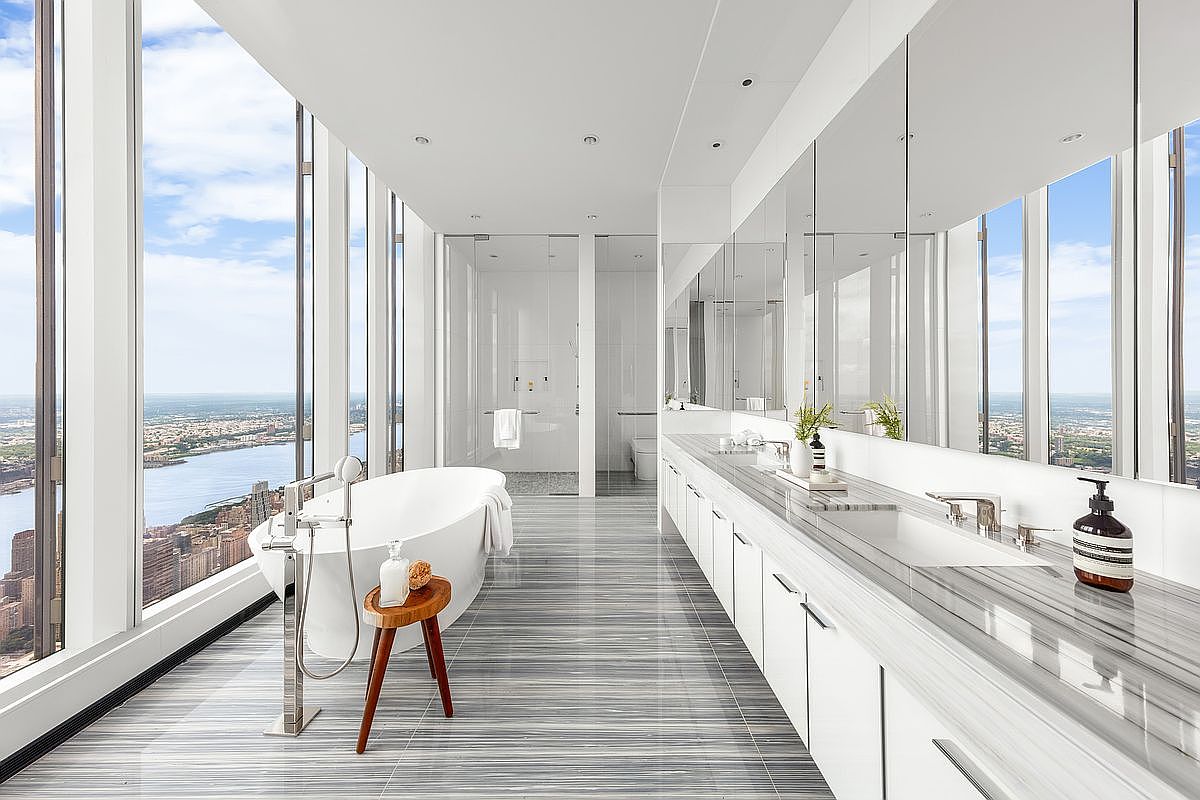This ultra-luxurious primary bathroom features floor-to-ceiling windows that offer a breathtaking, panoramic view of the city skyline and river. The space is defined by its sleek, minimalist aesthetic, showcasing a freestanding soaking tub, a long marble-topped double vanity, and polished grey-veined marble flooring. The clean lines and expansive glass create an airy, sophisticated atmosphere that emphasizes high-end urban living.