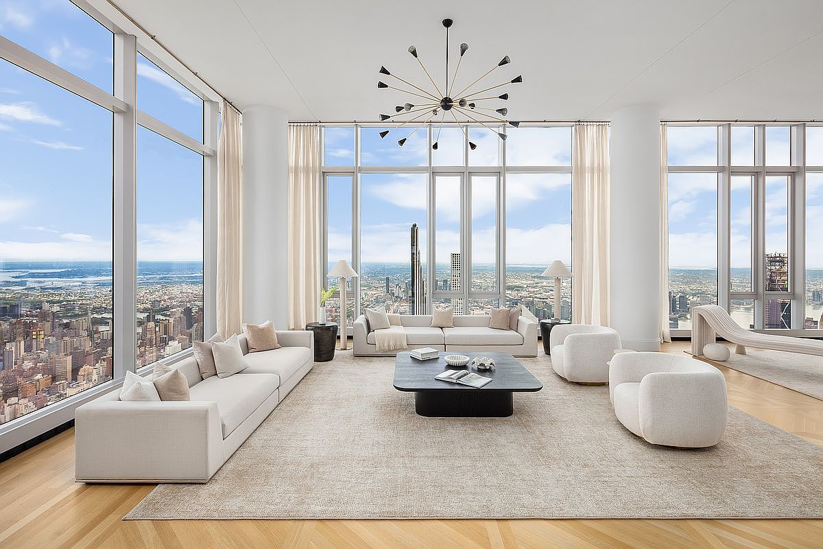 This expansive, high-floor living room features floor-to-ceiling windows that offer breathtaking, panoramic views of the city skyline and river. The space is furnished with plush, neutral-toned sofas and armchairs arranged around a sleek, dark coffee table, all set upon a large, textured area rug. A striking, modern sputnik-style chandelier serves as the focal point, enhancing the sophisticated and airy atmosphere of this luxury residence.