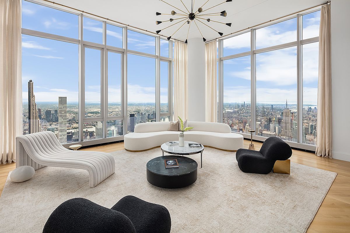 This luxurious, high-floor living room features floor-to-ceiling windows that offer a breathtaking panoramic view of the New York City skyline. The space is furnished with a sophisticated, curved white sofa, a modern black marble coffee table, and sculptural black accent chairs, all arranged on a plush, neutral-toned area rug. A striking, multi-armed sputnik-style chandelier serves as a dramatic focal point, enhancing the room's elegant and contemporary aesthetic.