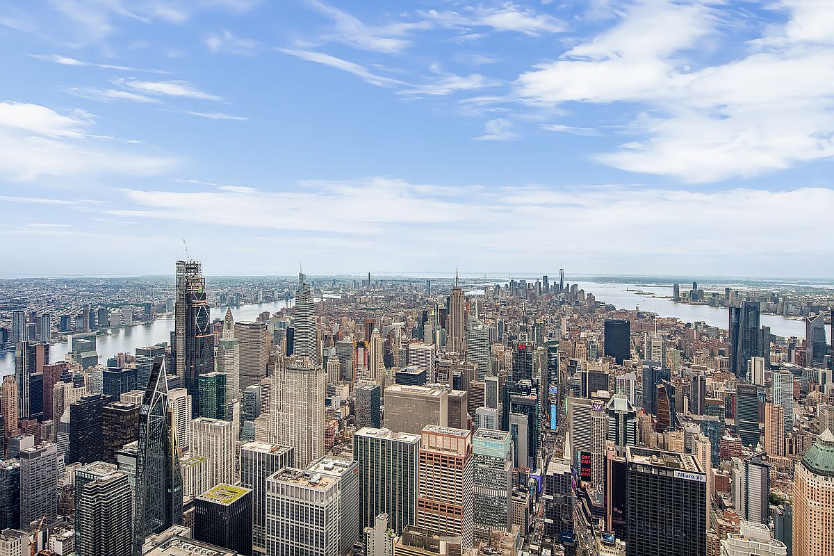 This high-angle aerial view captures the dense, iconic skyline of Manhattan, showcasing a vast array of skyscrapers and urban architecture. The perspective highlights the city's verticality and density, with the Hudson River visible in the distance under a bright, partly cloudy sky. It provides a grand, cinematic sense of scale, emphasizing the prime location and metropolitan lifestyle associated with the area.
