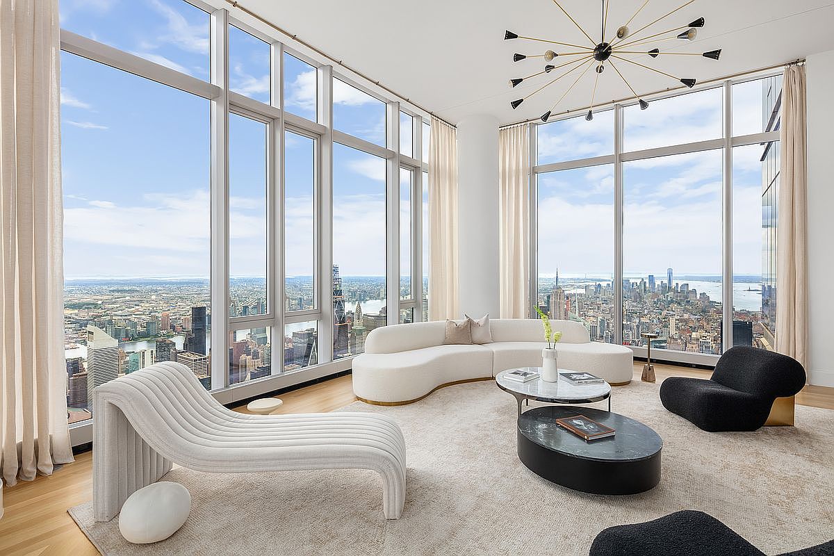 This sophisticated, high-rise living room features floor-to-ceiling windows that offer a breathtaking, panoramic view of the New York City skyline. The space is furnished with a contemporary, curved white sofa, a sculptural chaise lounge, and a modern black coffee table, all set upon a plush, neutral-toned area rug. A striking, multi-armed sputnik-style chandelier hangs from the ceiling, adding a touch of modern elegance to this bright and airy luxury apartment.