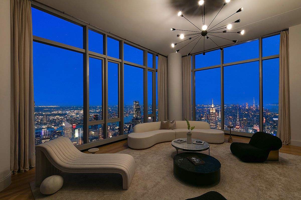 This sophisticated, high-rise living room features floor-to-ceiling windows that offer a breathtaking panoramic view of the New York City skyline at dusk. The space is furnished with a contemporary curved sofa, a sleek chaise lounge, and a dark, modern armchair, all arranged on a plush area rug around a minimalist coffee table. A striking sputnik-style chandelier hangs from the ceiling, adding a touch of mid-century modern elegance to the luxurious, open-concept interior.