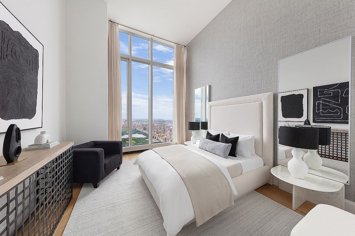 This sophisticated primary bedroom features a high-end, contemporary design with a plush upholstered bed, a sleek black armchair, and a unique metal-grid console table. The room is anchored by a large, floor-to-ceiling window that offers a breathtaking, elevated view of Central Park and the Manhattan skyline. The neutral color palette, textured wallpaper, and minimalist art create a serene and luxurious atmosphere.