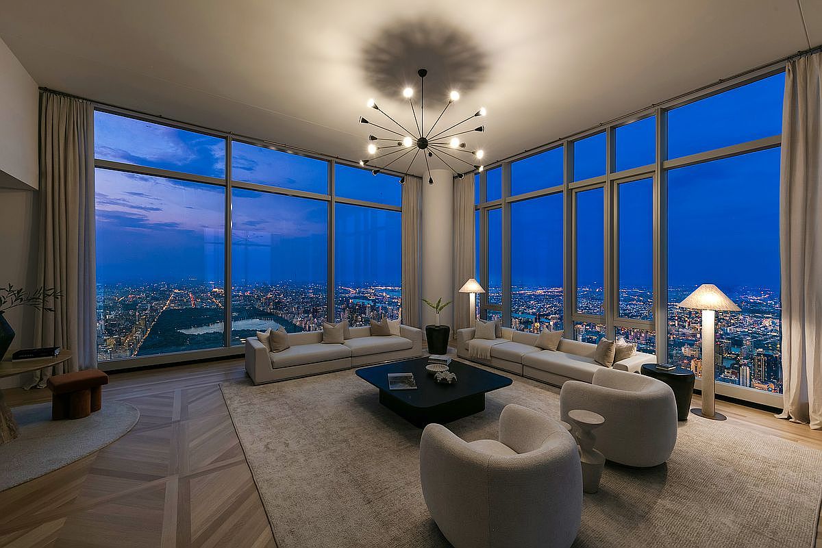 This sophisticated, high-rise living room features floor-to-ceiling windows that offer a breathtaking panoramic view of a sprawling city skyline at dusk. The interior is elegantly furnished with plush, neutral-toned sofas and armchairs arranged around a sleek, dark coffee table, all anchored by a large, textured area rug. A striking, modern sputnik-style chandelier serves as the focal point, casting a warm glow that complements the ambient twilight outside.