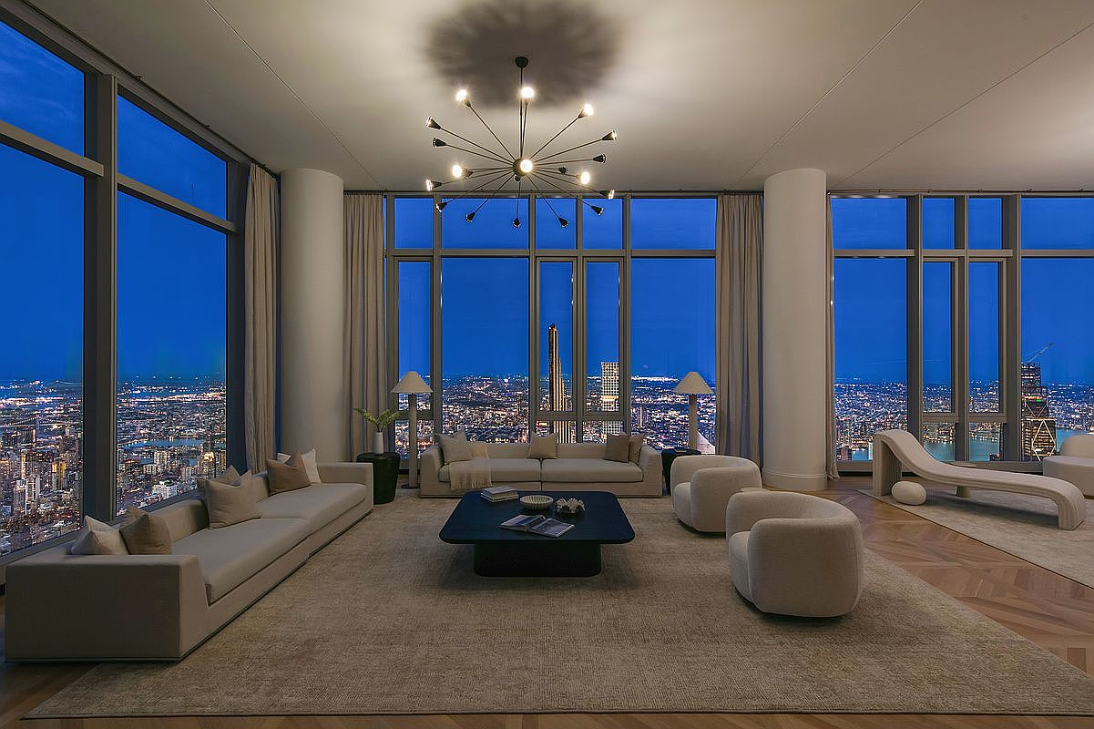 This expansive, high-end living room features floor-to-ceiling windows that offer a breathtaking, panoramic view of a sprawling city skyline at twilight. The interior is elegantly furnished with a long, neutral-toned sofa, a minimalist black coffee table, and plush, rounded accent chairs, all arranged on a large, textured area rug. A striking, modern sputnik-style chandelier serves as the centerpiece, casting a sophisticated glow over the open-concept space, which exudes luxury and contemporary refinement.
