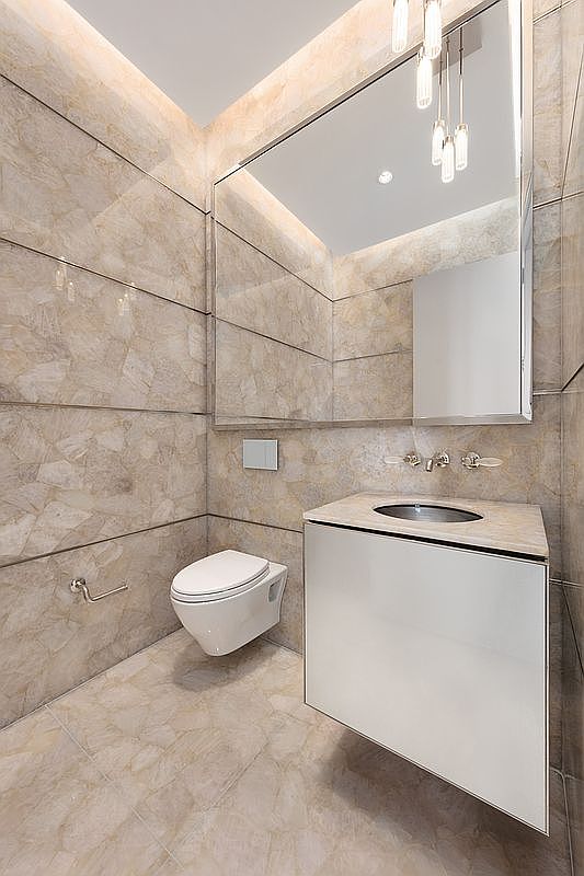 This elegant powder room features striking, large-format stone tiles with a warm, marbled texture covering the walls and floor. A sleek, wall-mounted white vanity with a circular sink and a minimalist wall-hung toilet create a clean, contemporary aesthetic. The space is elevated by a large, frameless mirror and sophisticated pendant lighting, offering a luxurious and refined atmosphere.