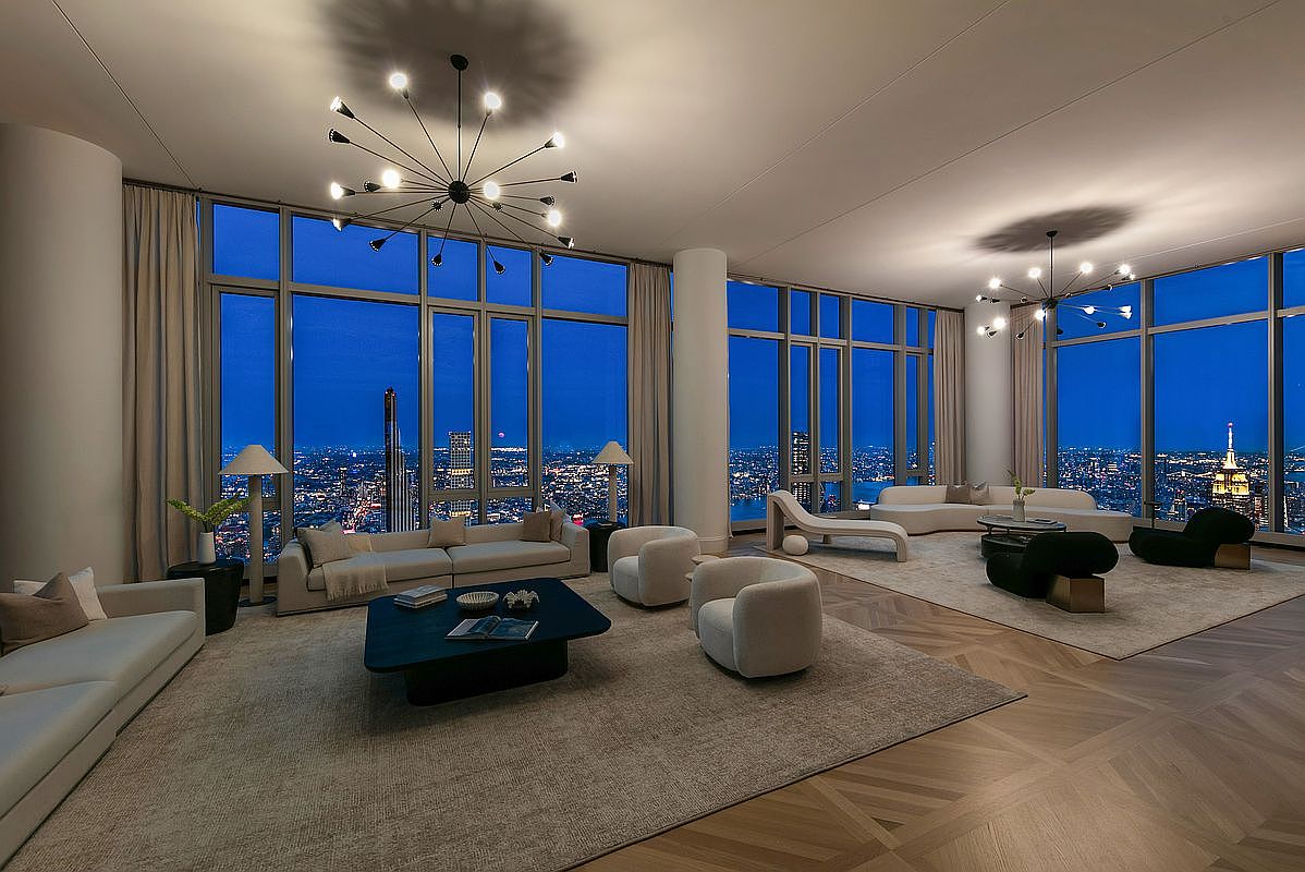 This expansive, high-end living room features floor-to-ceiling windows that offer a breathtaking panoramic view of the city skyline at twilight. The space is elegantly furnished with multiple curved sofas, modern armchairs, and minimalist coffee tables, all arranged on large, neutral-toned area rugs over herringbone wood flooring. Two striking, multi-armed sputnik-style chandeliers serve as the focal points, casting a warm, sophisticated glow across the luxurious, open-concept interior.