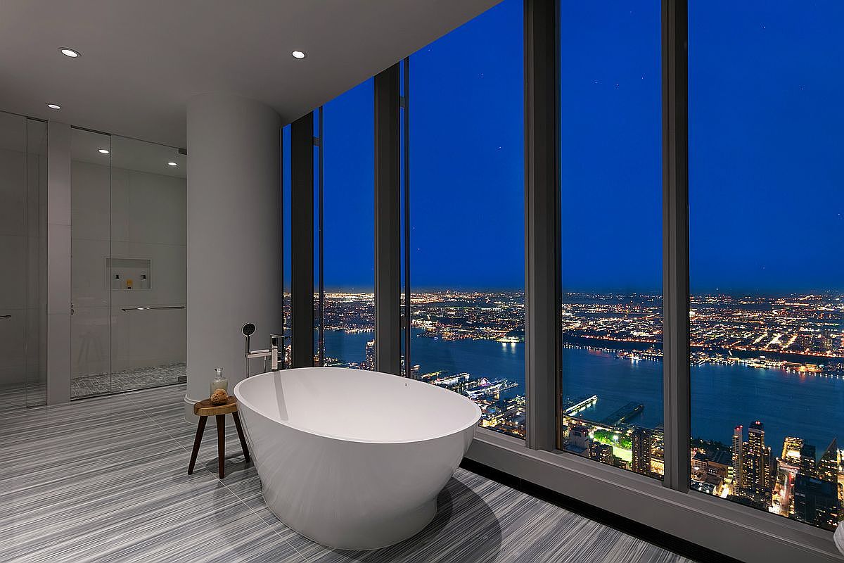 This luxurious primary bathroom features a sleek, freestanding white soaking tub positioned perfectly against floor-to-ceiling windows that offer a breathtaking, panoramic night view of the city skyline and river. The room is finished with sophisticated grey-toned striped marble flooring and includes a minimalist glass-enclosed shower area. The overall aesthetic is ultra-modern, high-end, and serene, emphasizing the dramatic urban backdrop.