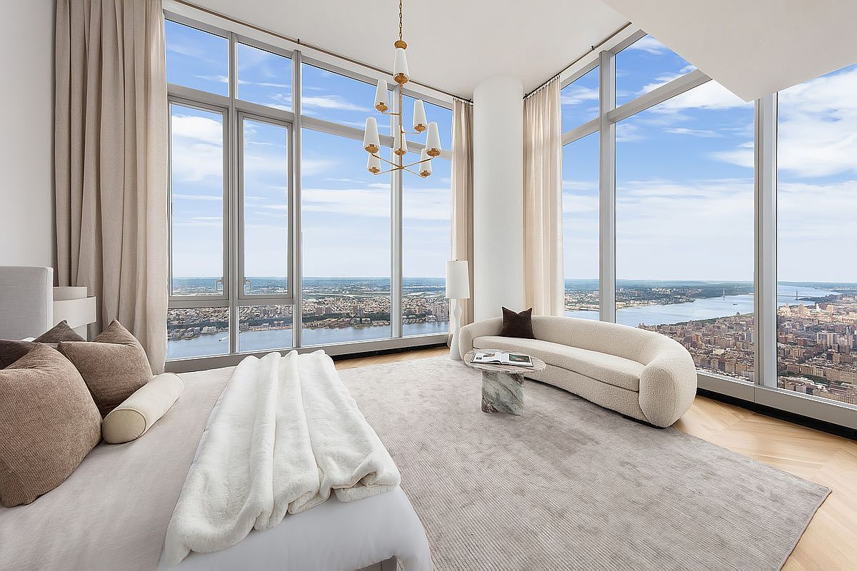 This luxurious primary bedroom features floor-to-ceiling windows that offer a breathtaking, panoramic view of the city skyline and river. The room is elegantly furnished with a plush, neutral-toned bed, a curved cream-colored sofa, and a sophisticated marble-based side table, all anchored by a soft, textured area rug. A modern, multi-armed chandelier hangs from the high ceiling, enhancing the airy and opulent atmosphere of this high-rise sanctuary.
