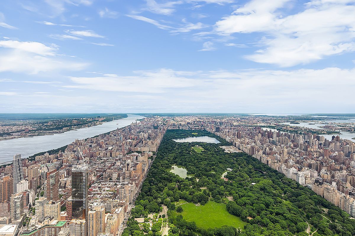 This high-altitude aerial view captures the iconic expanse of Central Park in New York City, flanked by dense urban development and skyscrapers on both sides. The perspective highlights the contrast between the lush green landscape of the park and the surrounding concrete jungle, with the Hudson River visible in the distance. The image provides a grand, cinematic sense of scale and location, emphasizing the prime real estate value of properties bordering this world-famous landmark.