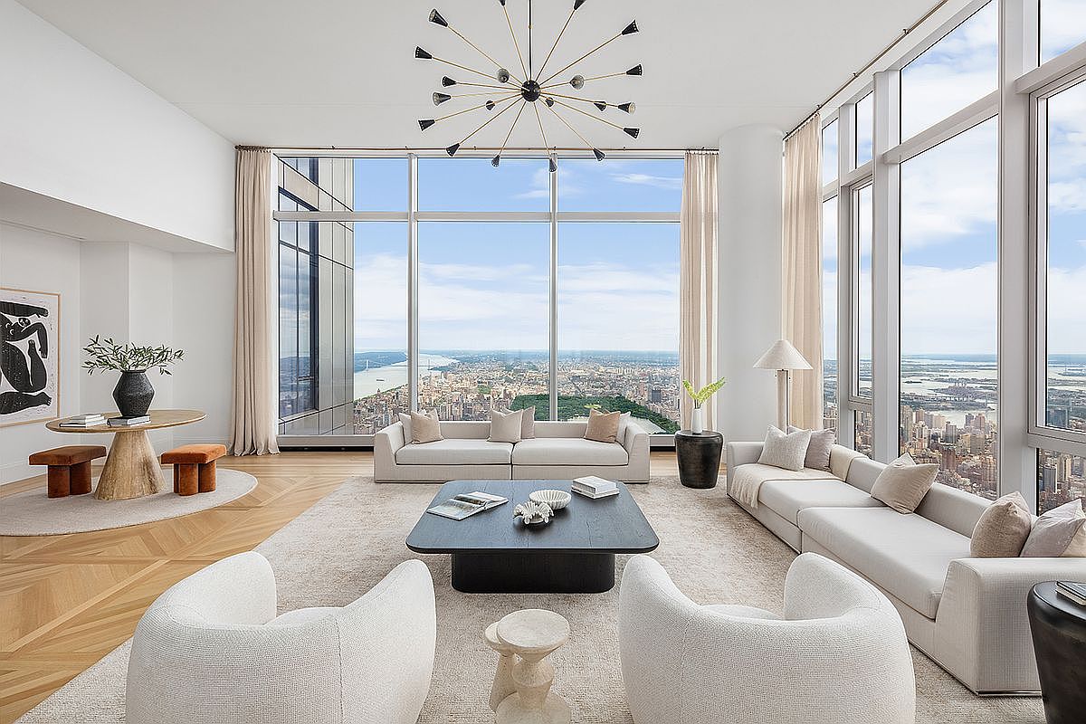 This expansive, ultra-luxury living room features floor-to-ceiling windows that offer a breathtaking, panoramic aerial view of the city skyline and river. The space is anchored by a sophisticated, neutral-toned seating arrangement, including plush white armchairs and a large, modern sectional, all centered around a sleek black coffee table. A striking, multi-armed sputnik-style chandelier hangs from the high ceiling, adding a touch of mid-century modern elegance to the bright, airy, and impeccably designed interior.