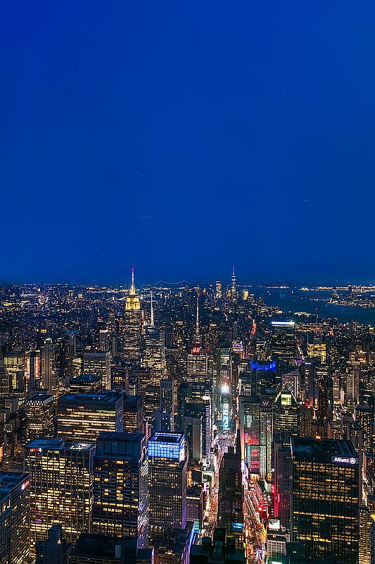 This stunning aerial view captures the iconic New York City skyline at twilight, showcasing a dense urban landscape illuminated by thousands of glowing windows and street lights. The perspective highlights the prominent Empire State Building standing tall amidst the architectural tapestry of skyscrapers, with the deep blue evening sky providing a dramatic backdrop. The scene conveys a sense of vibrant metropolitan energy and high-end urban living.