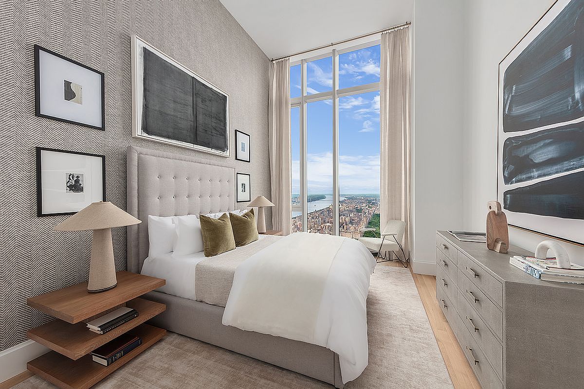 This sophisticated primary bedroom features a high-ceiling design with floor-to-ceiling windows that offer a breathtaking, expansive view of the city skyline and river. The room is elegantly appointed with a tufted upholstered bed, textured wallpaper, and a minimalist dresser, creating a serene and luxurious atmosphere. The perspective is captured from a slightly elevated angle, emphasizing the height of the room and the dramatic urban backdrop.