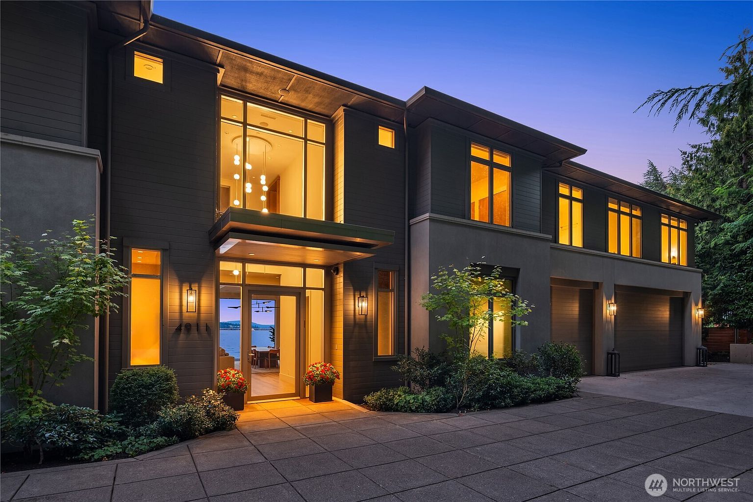 This striking modern luxury home features a dark, multi-textured facade with clean lines and expansive floor-to-ceiling windows that create a dramatic, illuminated entryway. The property includes a paved driveway leading to a multi-car garage, complemented by tasteful landscaping and a warm, inviting glow from the interior lighting at dusk. The perspective is a wide-angle, eye-level shot that emphasizes the home's architectural sophistication and its seamless integration with the surrounding environment.