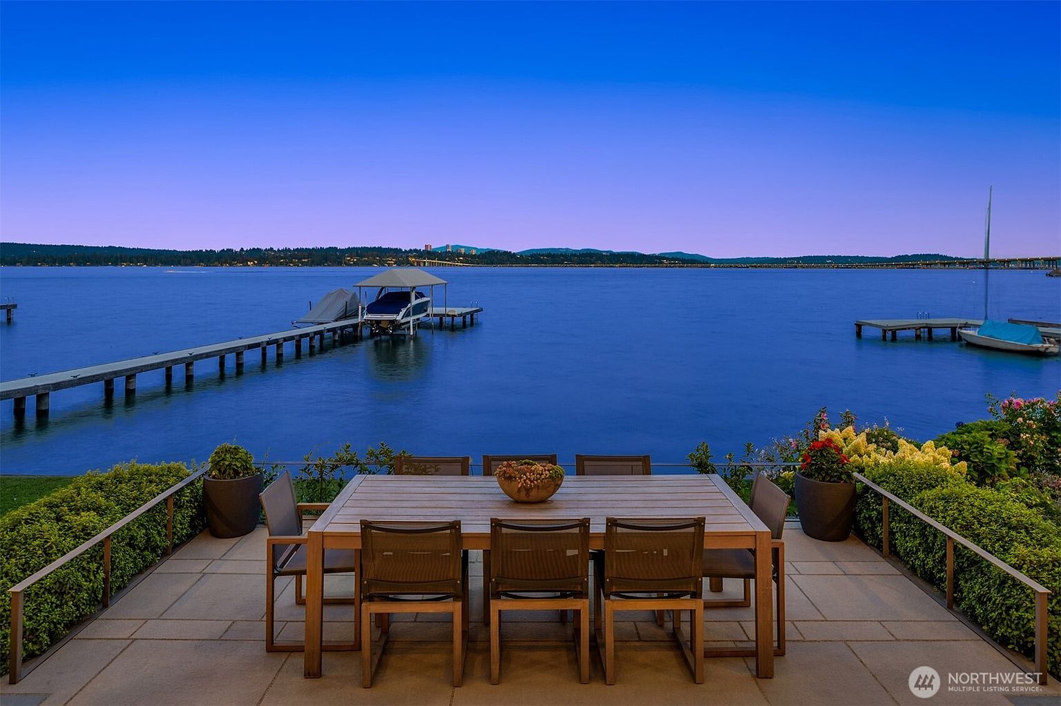 This serene outdoor patio features a large, modern wooden dining table set against a breathtaking panoramic view of a calm lake at dusk. The scene includes a private dock with a boat lift and a sailboat, framed by lush greenery and potted plants. The perspective is a wide, eye-level shot that emphasizes the tranquil waterfront lifestyle and the seamless transition between the home's living space and the natural surroundings.