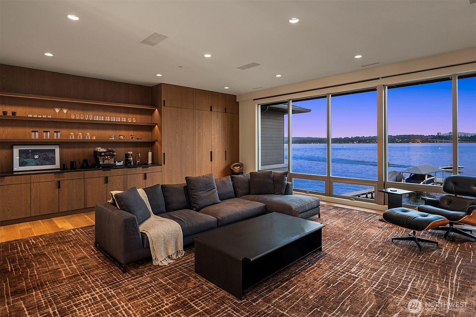 This sophisticated living room features a large, dark grey sectional sofa and a matching coffee table, set against a backdrop of custom wood cabinetry with integrated shelving. A floor-to-ceiling window wall provides a stunning, panoramic view of the lake, creating a seamless connection between the interior and the waterfront. The space is anchored by a textured, patterned area rug and accented with a classic lounge chair, exuding a modern, luxurious, and tranquil atmosphere.