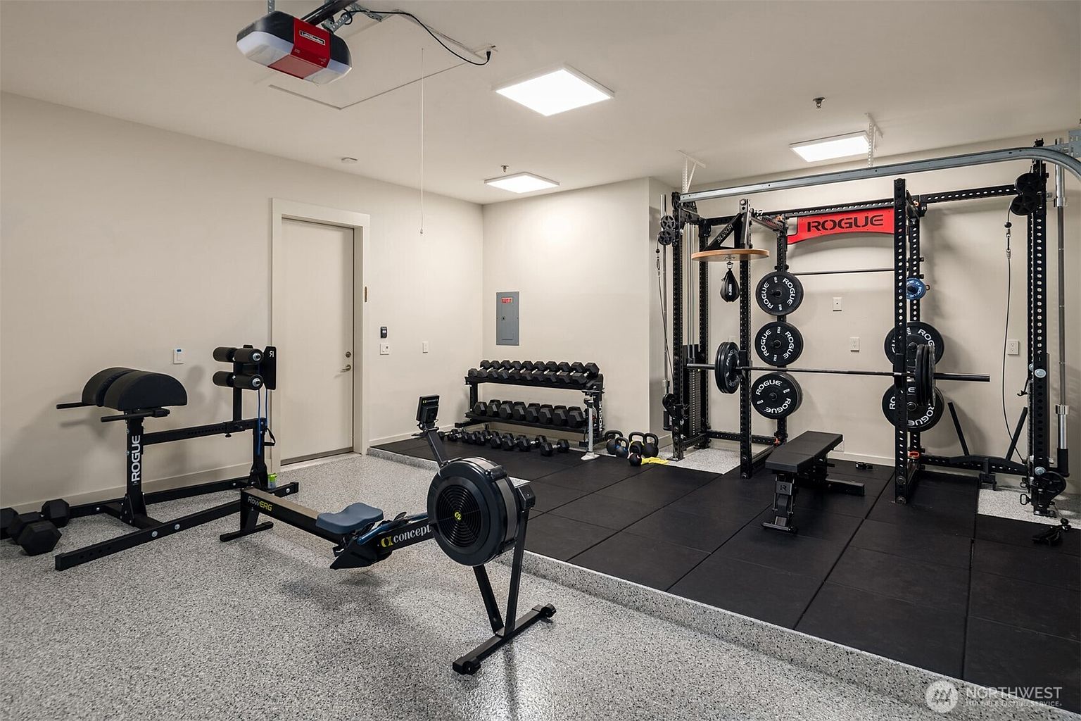 This well-equipped home gym is located within a garage space, featuring durable rubber flooring and high-quality Rogue fitness equipment. The setup includes a power rack with a barbell, a Concept2 rowing machine, a glute-ham developer, and a comprehensive rack of dumbbells. The space is brightly lit with modern LED panel lighting, creating an inviting and professional atmosphere for a home workout.