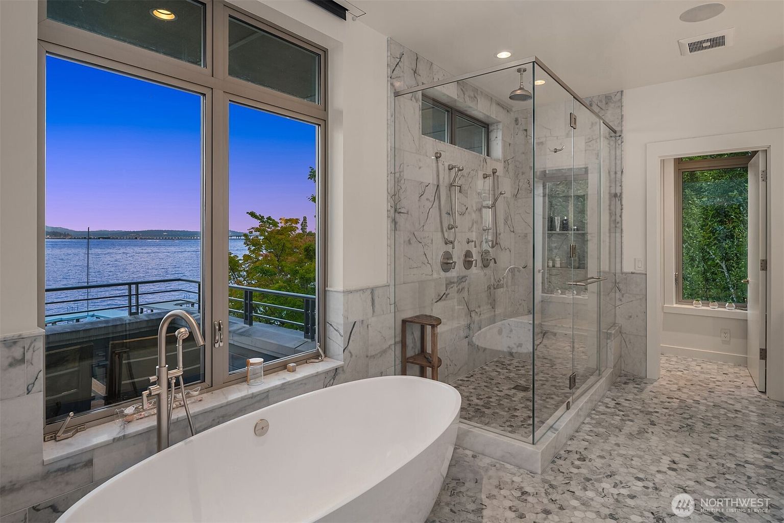 This luxurious primary bathroom features a sleek, freestanding soaking tub positioned perfectly in front of a large window that offers a stunning, panoramic view of the water. Adjacent to the tub is a spacious, glass-enclosed walk-in shower with marble tiling and multiple showerheads, creating a spa-like atmosphere. The room is finished with elegant marble flooring and high-end fixtures, emphasizing a sophisticated and serene aesthetic.