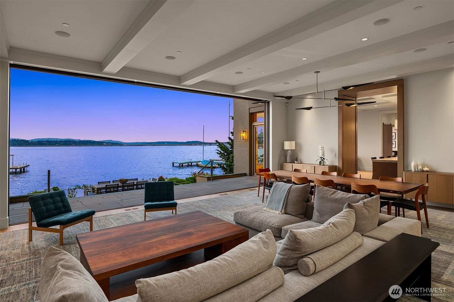 This expansive, modern living room features an open-concept design that seamlessly transitions to an outdoor deck through a massive, retractable glass wall overlooking a serene lake. The space is furnished with a large, plush sectional sofa, a rich wooden coffee table, and contemporary accent chairs, all anchored by a textured area rug. The room is illuminated by recessed lighting and a sleek, minimalist chandelier, creating an elegant and airy atmosphere perfect for luxury waterfront living.