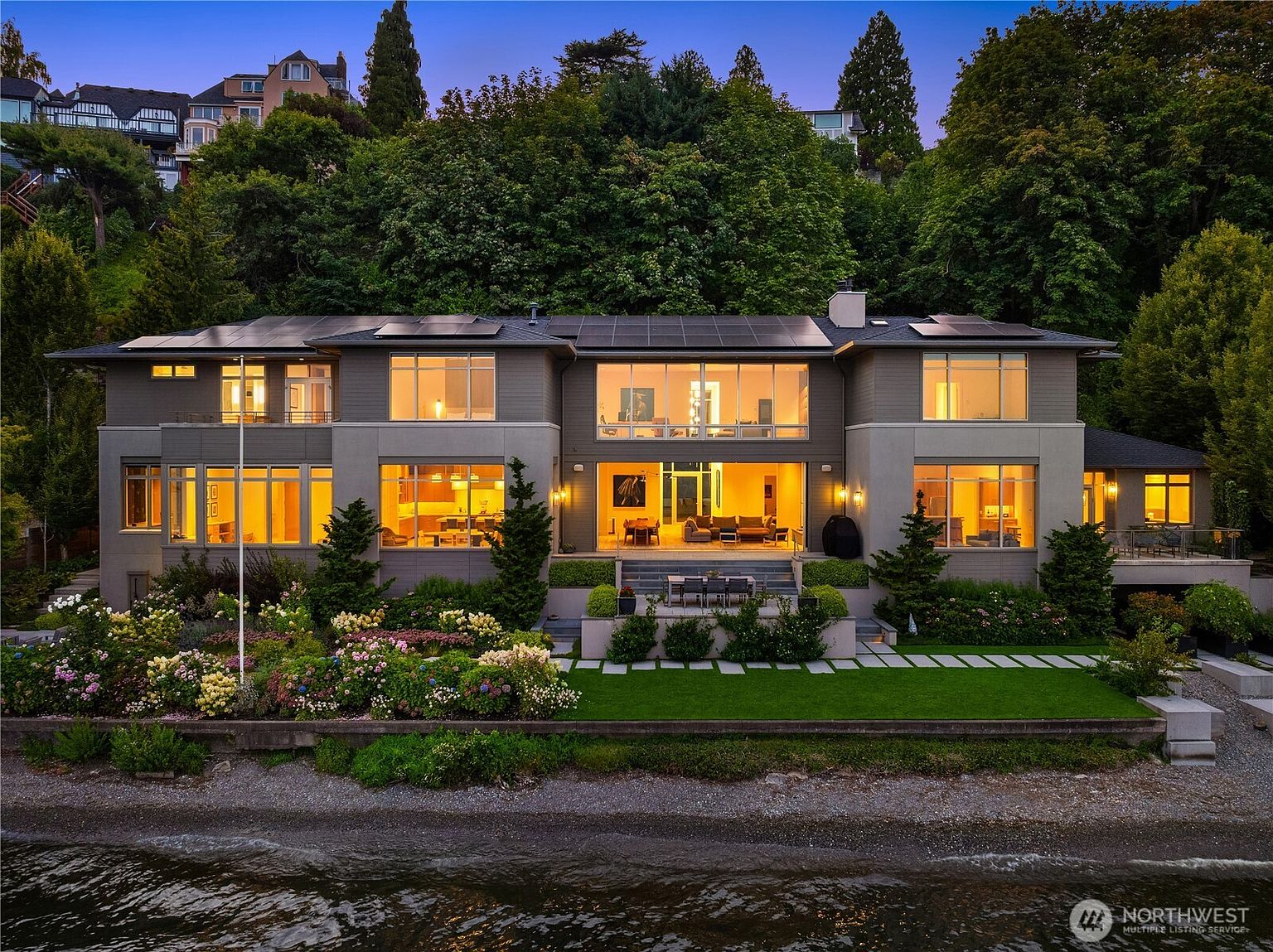 This stunning waterfront estate features a contemporary architectural design with expansive floor-to-ceiling windows that offer panoramic views of the water. The multi-level home is nestled against a lush, forested hillside and includes a meticulously landscaped garden, a stone patio, and integrated solar panels on the roof. The warm interior lighting creates an inviting atmosphere, highlighting the seamless transition between the luxurious living spaces and the serene outdoor environment.
