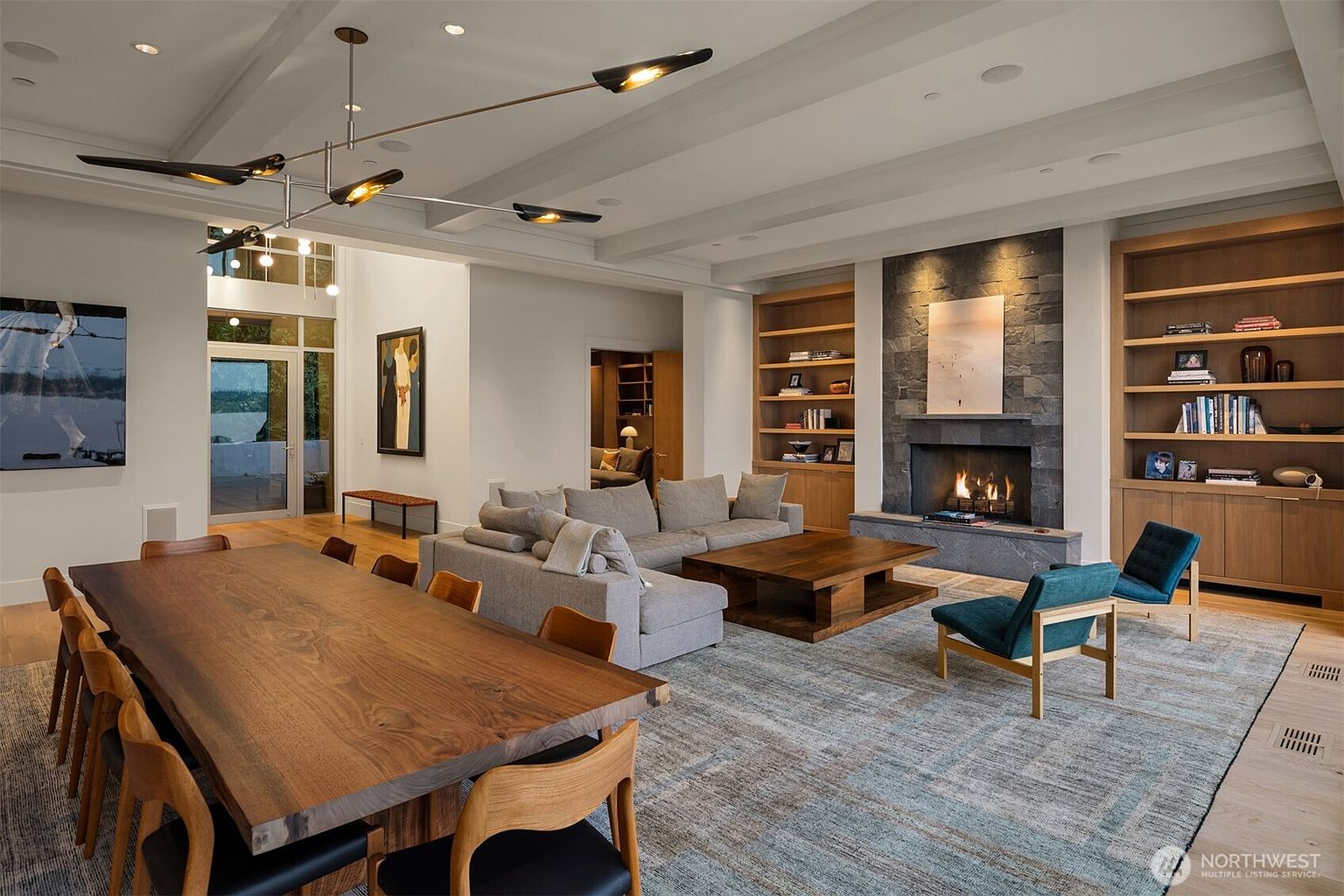 This expansive open-concept living and dining area features a sophisticated blend of modern design and warm, natural materials. A striking live-edge wood dining table anchors the foreground, while the living space centers around a dramatic floor-to-ceiling stone fireplace flanked by custom built-in shelving. The room is bathed in natural light from large windows and is accented by a contemporary chandelier, creating an inviting and luxurious atmosphere.