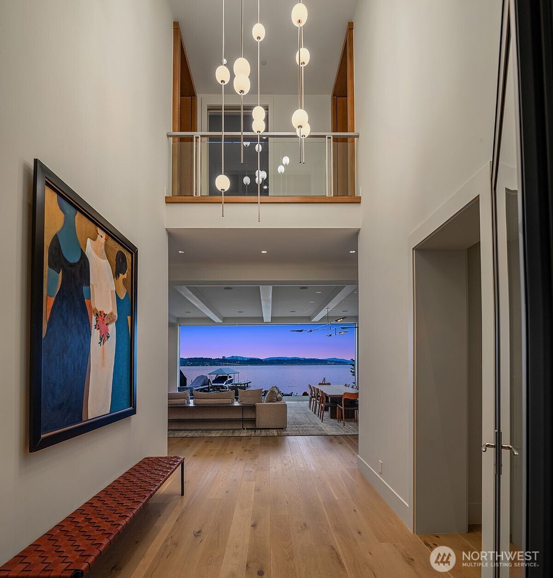 This striking entryway features a dramatic double-height ceiling with a cascading modern chandelier that draws the eye upward toward an open loft landing. The perspective looks through the hallway, past a large abstract painting and a woven bench, toward a breathtaking, unobstructed view of the water and sunset through floor-to-ceiling glass walls. The clean, minimalist aesthetic and warm wood flooring create a sophisticated and inviting atmosphere that seamlessly blends indoor luxury with the natural landscape.