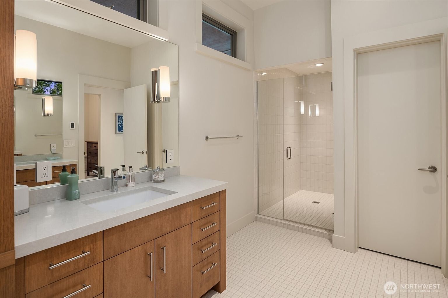 This modern primary bathroom features a sleek wood-toned vanity with a light-colored stone countertop and a large, frameless mirror. To the right, a spacious walk-in shower with white subway tile and a glass door creates a clean, minimalist aesthetic. The room is brightened by natural light from a high transom window and contemporary wall sconces, offering a polished and functional design.