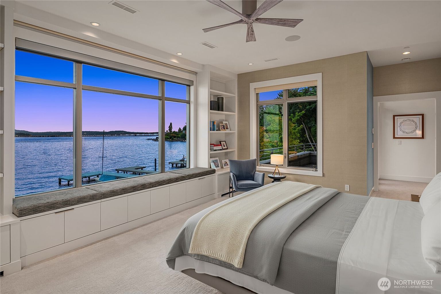 This luxurious primary bedroom features a stunning floor-to-ceiling window wall that offers a panoramic view of the lake, complete with a built-in window seat and storage cabinetry. The room is elegantly appointed with a plush bed, a cozy reading nook with a blue armchair, and built-in shelving, all set against a neutral, sophisticated color palette. The perspective captures the serene atmosphere of the space, highlighting the seamless integration of indoor comfort and breathtaking waterfront scenery.
