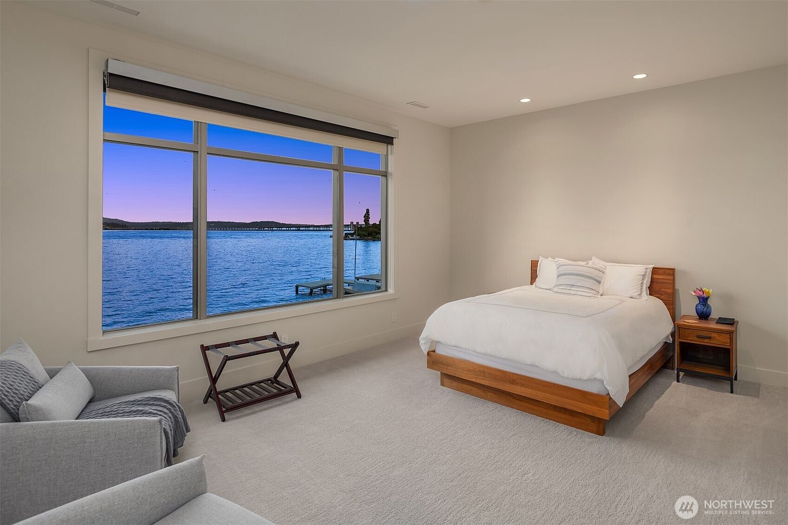 This serene primary bedroom features a minimalist aesthetic with a platform bed as the focal point, complemented by a wooden nightstand and a cozy armchair in the foreground. A large, expansive window offers a breathtaking, unobstructed view of the lake, bathing the room in natural light and creating a tranquil atmosphere. The neutral color palette and clean lines emphasize a modern, sophisticated design perfect for a waterfront retreat.