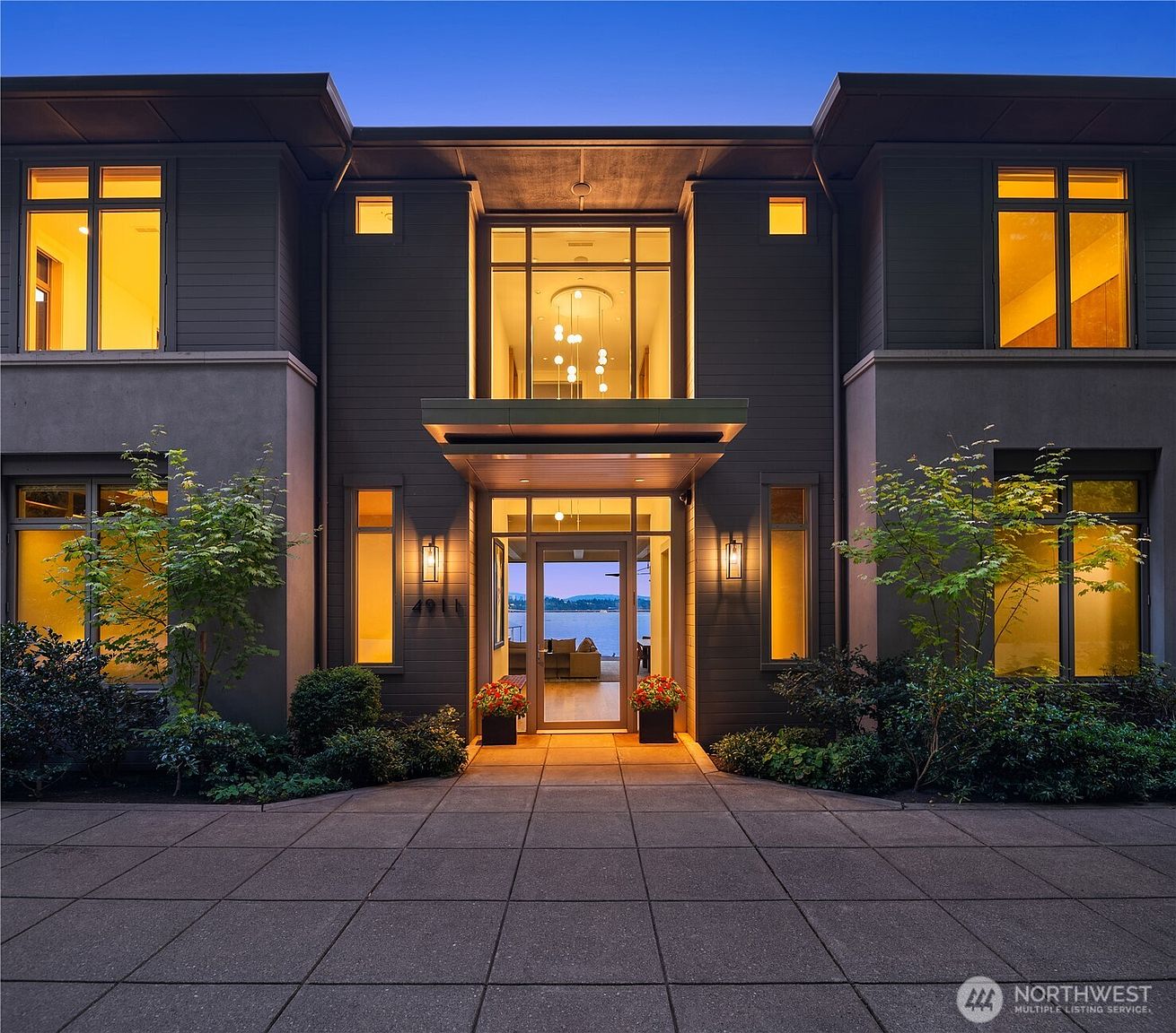 This striking modern residence features a symmetrical, dark-toned facade with clean lines and expansive glass windows that glow warmly against the twilight sky. A prominent central entryway is framed by a cantilevered overhang and a tall, multi-story glass panel, creating a sophisticated and inviting focal point. The paved driveway leads directly to the front door, where a glimpse through the house reveals a serene waterfront view, emphasizing the property's premium location.