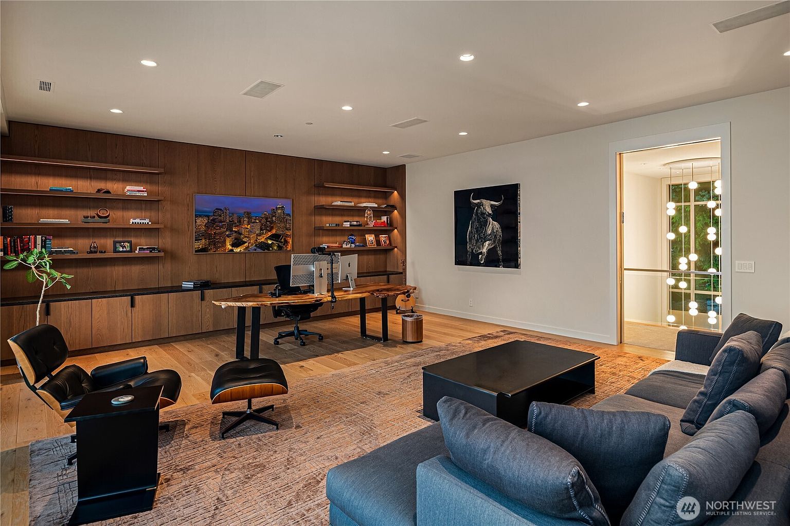 This sophisticated home office features a striking live-edge wood desk as the centerpiece, set against a wall of custom floor-to-ceiling wood shelving and cabinetry. The room includes a comfortable lounge area with a dark grey sofa and an iconic Eames-style lounge chair, all anchored by a textured area rug. A large bull portrait adds a bold artistic statement, while the open doorway reveals a modern staircase illuminated by a unique, cascading globe chandelier.