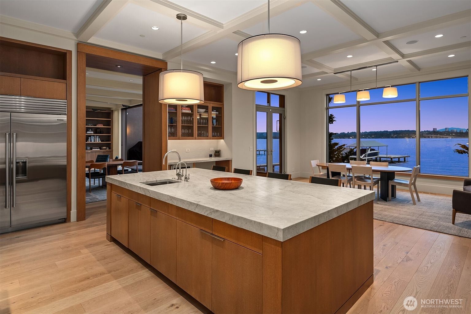 This sophisticated, modern kitchen features a large central island with a light-toned stone countertop and warm wood cabinetry, creating a seamless blend of natural textures. The open-concept design flows into a dining area with expansive floor-to-ceiling windows that frame a stunning waterfront view. The space is illuminated by elegant pendant lighting and recessed ceiling lights, emphasizing the high-end finishes and airy, luxurious atmosphere.