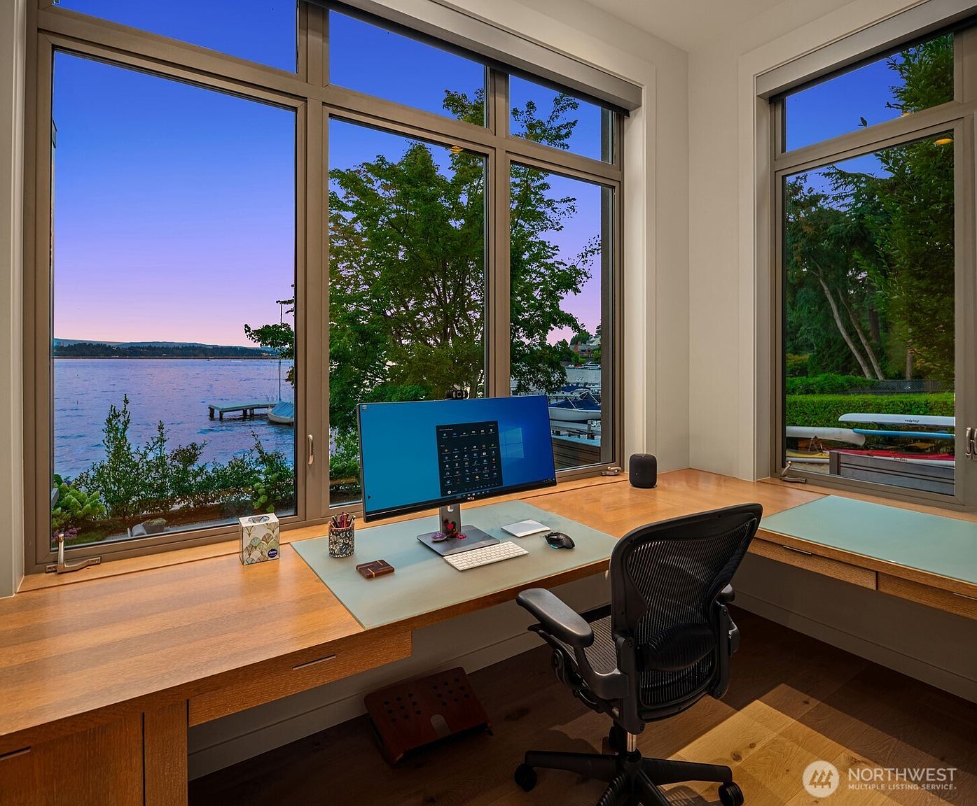 This sophisticated home office features a custom-built, wrap-around wooden desk positioned perfectly to take advantage of expansive, floor-to-ceiling windows overlooking a serene waterfront. The room is furnished with a high-end ergonomic mesh office chair and a sleek, modern computer setup, creating a productive yet tranquil workspace. The clean lines, warm wood tones, and breathtaking natural views combine to offer a premium, professional atmosphere.