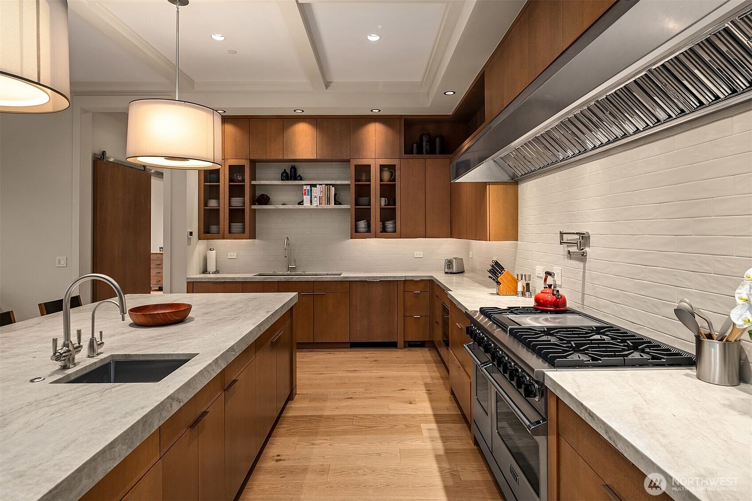 This sophisticated, modern kitchen features warm wood cabinetry, expansive light-toned stone countertops, and a large central island with an integrated sink. The space is illuminated by elegant pendant lighting and recessed ceiling lights, highlighting the high-end stainless steel range and professional-grade hood. The open-concept design, complemented by light hardwood flooring, creates a seamless and inviting atmosphere perfect for both cooking and entertaining.
