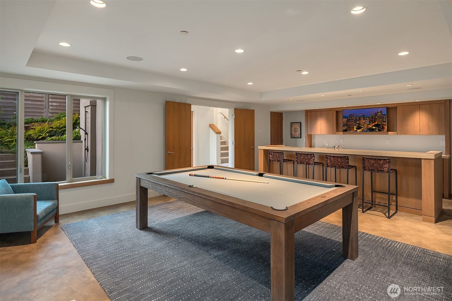 This spacious, modern basement recreation room features a large wooden pool table as the centerpiece, set upon a textured gray area rug. To the right, a built-in wet bar with wooden cabinetry, a wall-mounted television, and four leather bar stools creates an ideal entertainment space. The room is illuminated by recessed lighting and includes a large window looking out onto a landscaped outdoor area, providing a bright and inviting atmosphere.