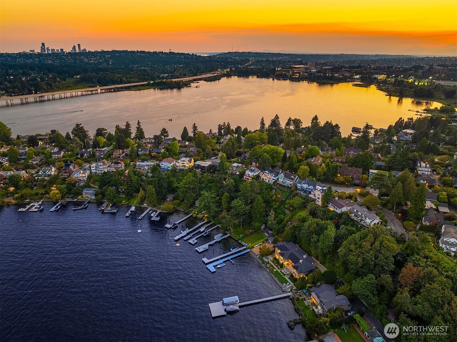 This stunning aerial perspective captures a luxurious waterfront property nestled along a lush, tree-lined shoreline at sunset. The scene features a private dock extending into the deep blue water, with a vibrant orange and yellow sky reflecting off the lake and the distant city skyline. The composition highlights the serene, upscale residential neighborhood and the seamless integration of the home with its natural surroundings.