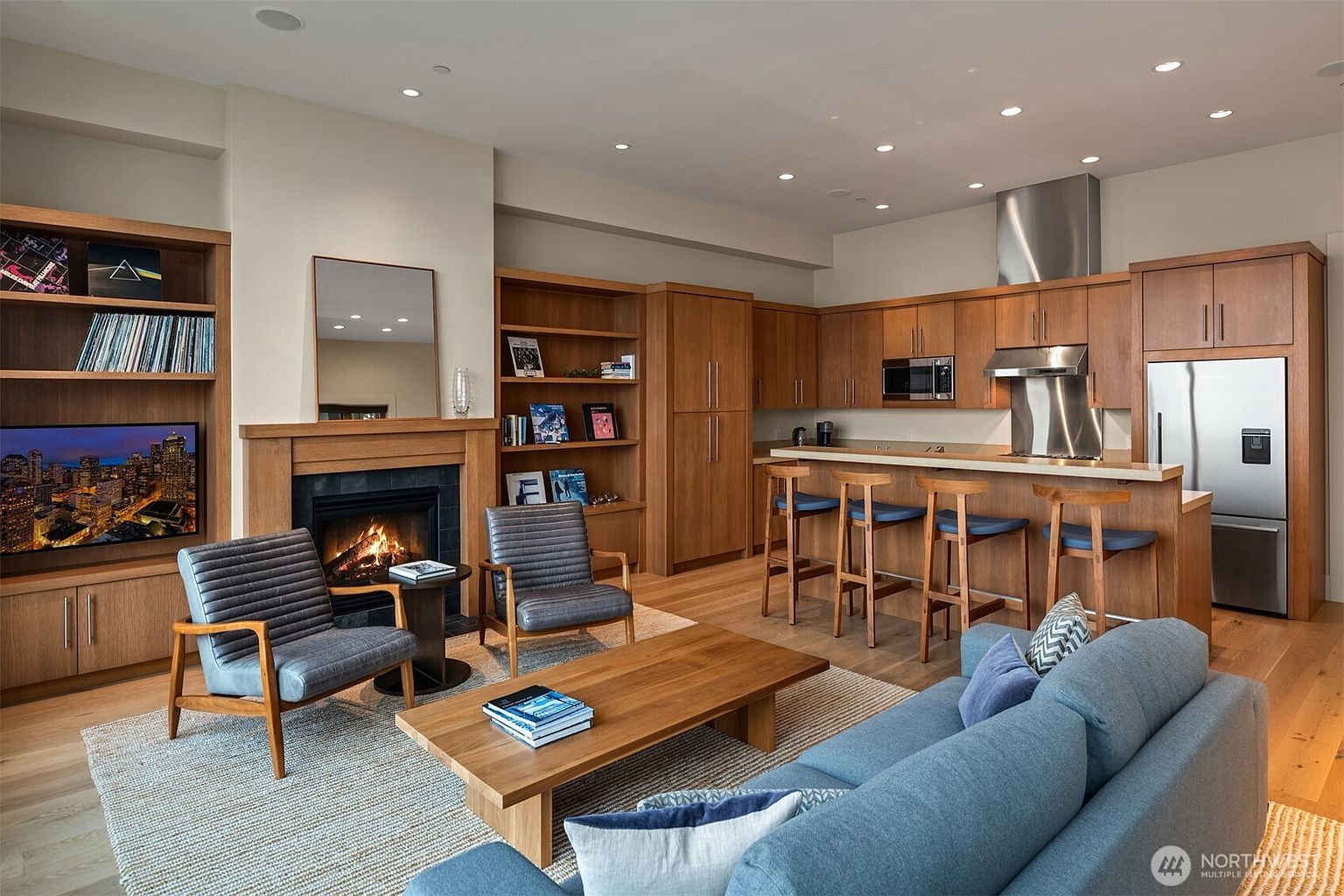 This open-concept living space features a warm, contemporary design with light hardwood floors and rich wood cabinetry. The room centers around a cozy fireplace with built-in shelving, flanked by two modern grey armchairs and a plush blue sofa, while the adjacent kitchen area includes a breakfast bar with four stools. The perspective captures a seamless flow between the relaxing living area and the functional kitchen, creating an inviting and cohesive home environment.