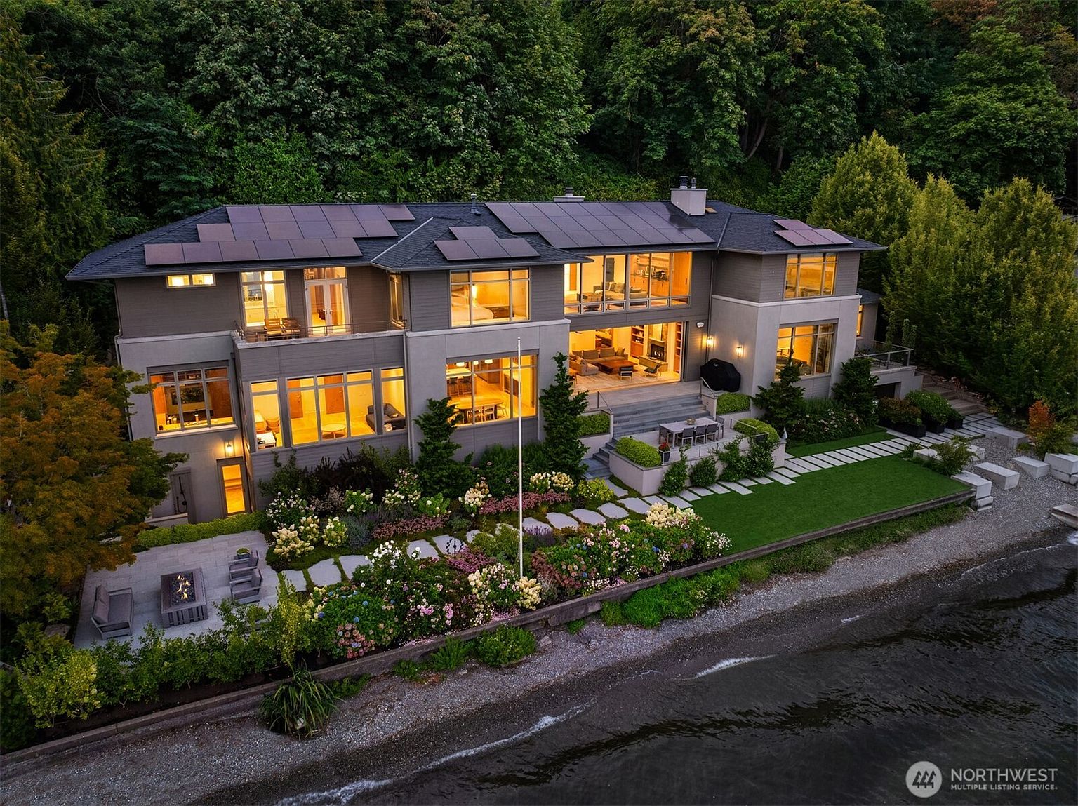 This stunning aerial view captures a luxurious, multi-story waterfront residence nestled against a lush, forested hillside. The home features a contemporary design with expansive floor-to-ceiling windows, solar panels on the roof, and a beautifully landscaped garden that transitions into a private stone patio and beach access. The warm interior lighting creates an inviting atmosphere, highlighting the seamless integration between the sophisticated architecture and the serene natural surroundings.