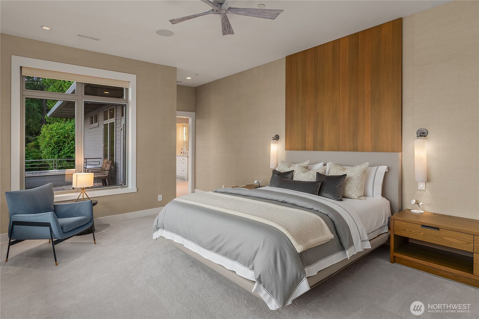 This elegant primary bedroom features a sophisticated neutral color palette, highlighted by a striking floor-to-ceiling wood accent wall behind the bed. The room is furnished with a plush, layered bed, a modern blue armchair, and sleek wooden nightstands, all set against soft carpeting. A large window provides ample natural light and a view of the outdoor deck, creating a serene and luxurious atmosphere.