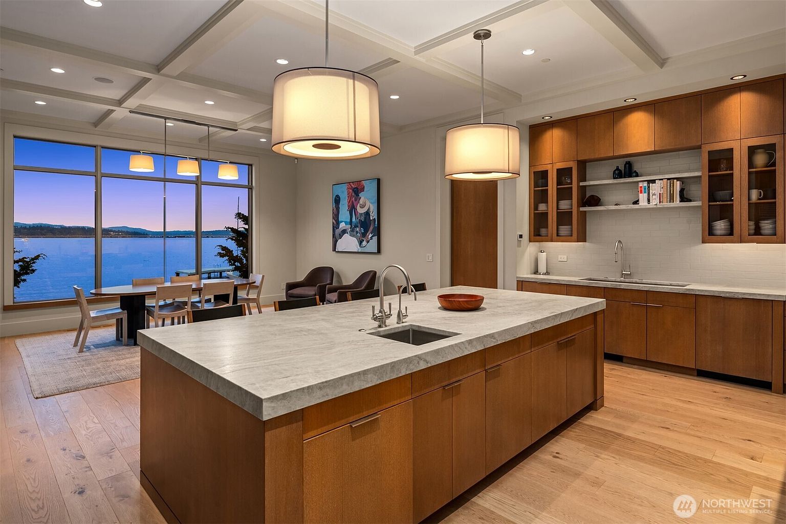 This sophisticated, open-concept kitchen features warm wood cabinetry, a large central island with a stone countertop, and a seamless transition to a dining area overlooking a serene waterfront view. The space is illuminated by modern pendant lighting and recessed ceiling lights, highlighting the clean lines and contemporary design. The expansive floor-to-ceiling windows provide a stunning, panoramic backdrop that enhances the airy and luxurious atmosphere of the home.